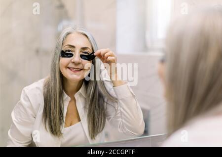 Gesichtsmaske und Anti-Falten-Therapie. Lächelnde ältere Frau, die vor dem Badezimmerspiegel steht und schwarze Augenflecken nach der Anstellung wieder anfärbt Stockfoto