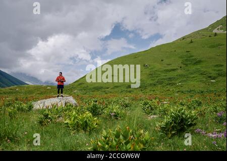 Junger Tourist mit Kamera steht auf dem großen Stein inmitten der grünen Hügel in den Bergen. Stockfoto