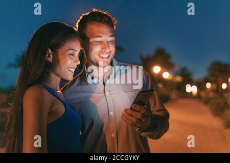 Glückliches Paar, das nachts auf der Stadtstraße mit dem Handy telefoniert hat, hat das Licht des Telefons beleuchtet und online nach einem Restaurant gesucht, um ausgehen zu können. Smartphone-Nutzer Stockfoto