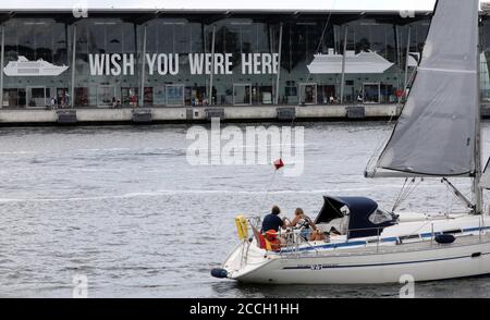 Rostock, Deutschland. August 2020. Ein Segelboot passiert den leeren Kreuzfahrtterminal, wo "WISH YOU WERE HERE" steht, während das Kreuzfahrtschiff "MS Europa 2" für einen kurzen Zwischenstopp gekommen ist. Die 'MS Europa 2' entschied sich jedoch, nicht anzudocken und verließ die Stadt sofort. Kreuzfahrtschiffe mit Passagieren legen aufgrund der Corona-Bedingungen derzeit nicht in Rostock an. Quelle: Bernd Wüstneck/dpa-Zentralbild/dpa/Alamy Live News Stockfoto