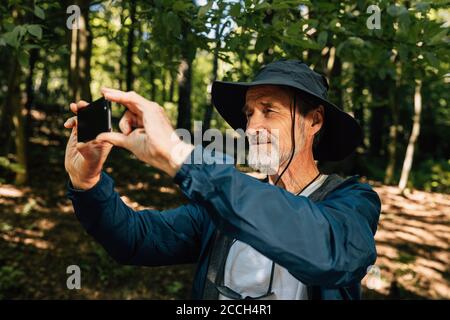 Seniortourist, der im Wald steht und fotografiert. Bärtiger Mann auf Wandertour, der Fotos auf seinem Handy machte. Stockfoto