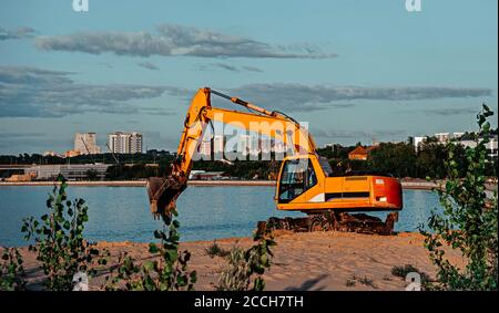 Orange Bagger arbeitet am Flussufer. Sand, Wasser und Bäume im Hintergrund. Stockfoto