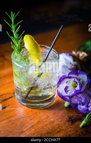 Ein elegant aussehender Craft-Cocktail aus rockigem Glas mit Zitronen- und Rosmaringarnitur sitzt neben einer Blume in einer eleganten, stilvollen, rustikalen Bar-Umgebung. Stockfoto