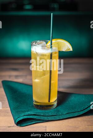 Ein gelber Cocktail in einem collins Glas sitzt auf einem Grüne Serviette auf einem grau gebeizten Holztisch Stockfoto