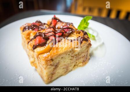 Ein weißer Teller mit einer Scheibe frisch gebackenem Brot Pudding mit Erdbeeren bedeckt und mit Schokolade beträufelt Stockfoto