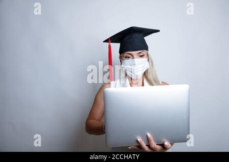 Eine schöne Absolventin in einer Schutzmaske auf ihrem Gesicht und einer Abschlusskappe hält einen Laptop in der Hand. Hochwertige Fotos Stockfoto