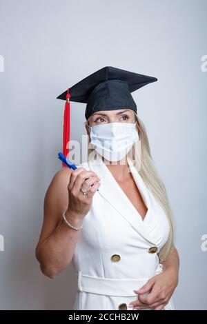 Eine schöne Absolventin, die eine medizinische Schutzmaske auf ihrem Gesicht und eine Abschlusskappe auf ihrem Kopf trägt, hält einen Stift in ihrer Hand. Hochwertige Fotos Stockfoto
