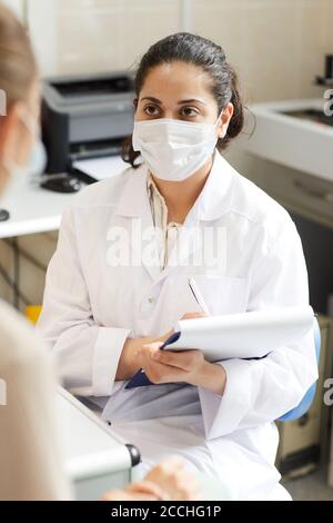 Weibliche Arzt in Maske und weißen Mantel Schreiben Rezept für Patient im Krankenhaus Stockfoto