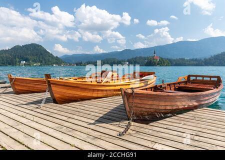 Nahaufnahme des slowenischen Sees Bled, mit einem hölzernen Pier und drei Reihenbooten, die unter einem blauen Sommerhimmel mit geschwollenen Wolken vertäut sind Stockfoto