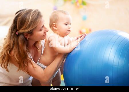 Seitenansicht der jungen kaukasischen Mutter hilft kleinen Baby trägt Windel üben Gymnastik Übungen zu Hause. Glückliche Frau Ausbildung Kind mit großen BL Stockfoto