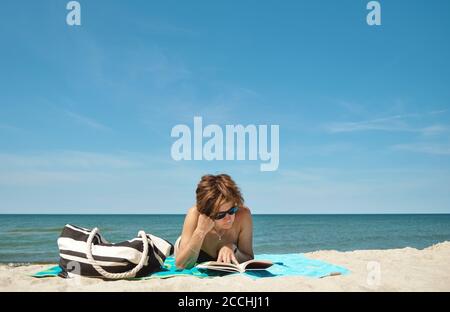 Attraktive Frau mittleren Alters kaukasischen in Bikini liegend auf der Strand und Lesen eines Buches am hellen Sommertag Stockfoto