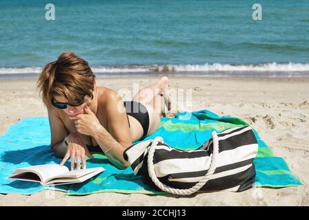 Kaukasische Frau mittleren Alters im Bikini am Strand liegend Lesen eines Buches über hellen Sommertag Stockfoto