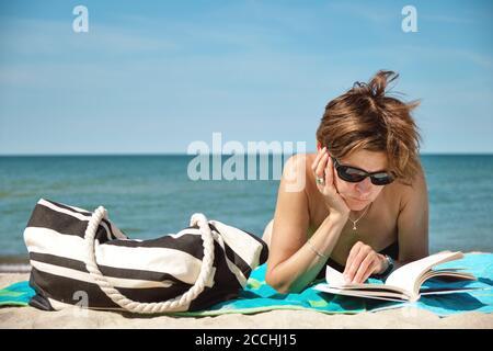 Nahaufnahme einer kaukasischen Frau mittleren Alters im Bikini, auf dem man sonnenbaden kann Der Strand und das Lesen eines Buches am hellen Sommertag Stockfoto