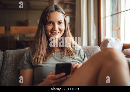Portrait der schönen jungen Frau hält Handy während des Sitzens Auf dem Sofa im Wohnzimmer Stockfoto