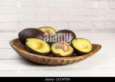 Reife braune Avocados ( hass - bilse Variety ) einige von ihnen halbiert in Holz geschnitzte Schüssel, auf weißen Brettern Schreibtisch, Backstein Wand Hintergrund Stockfoto