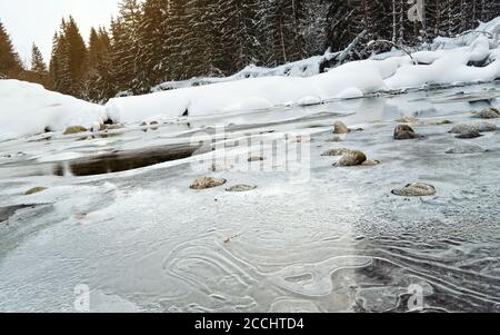 Morgen auf gefrorenem Fluss im Winter, Low-Angle-Foto, Fokus auf Eis-Muster und Steine, mit Nadelbäumen und aufgehenden Sonne in der Ferne Stockfoto