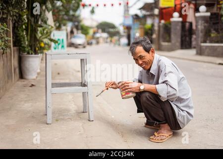 Hoi an / Vietnam - 18. Januar 2020: Vietnamesischer Mann lächelt, während er Holzbank mit Pinsel auf der Straße malt Stockfoto