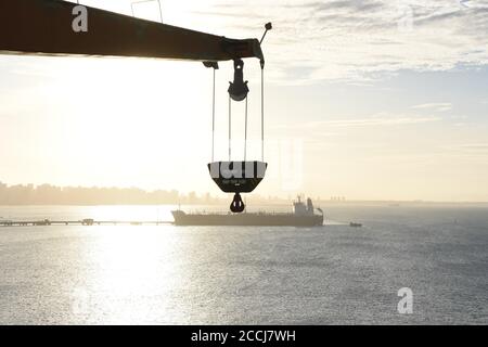 Blick auf die Silhouette eines Tankschiffes unter der Sonne entlang des Entladerohres, das scheinbar am Ausleger und Haken des Frachtschiffes hängt. Stockfoto