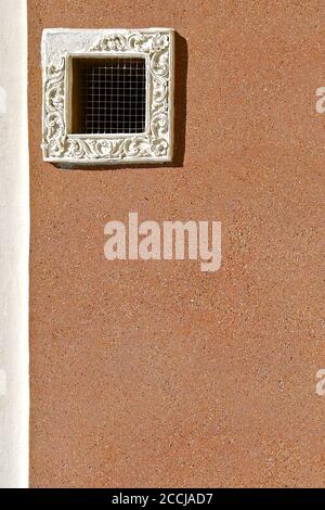 Kleiner quadratischer Lüftungsschlitz mit Hühnerdrahtgitter und einem verzierten weißen Gipsfensterrahmen. Isoliert auf einer sandigen farbigen Kieswand gestrichelt. Stockfoto