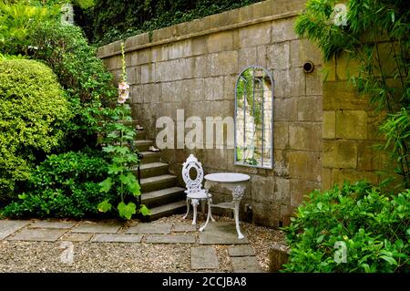 Ein Hof Landgarten mit Steinmauern, Stufen und markierten Wegen. Mit weißen schmiedeeisernen Terrassenmöbeln, einem Spiegel im georgianischen Stil und üppigen Bäumen, Stockfoto