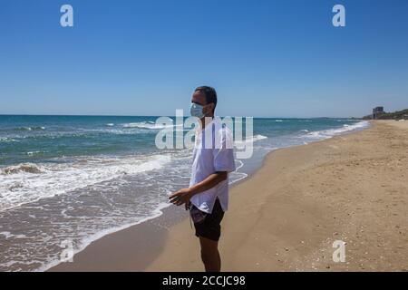 Mann mit medizinischer Maske gegen Coronavirus / covid 19, zu Fuß am Strand während der Pandemie, Quarantäne.sicheres Reisekonzept. Stockfoto