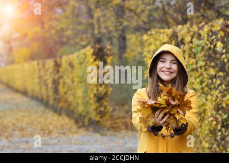 Teenagermädchen in einem gelben Kapuzenmantel hält die Blätter In ihren Händen Herbstsaison Stockfoto