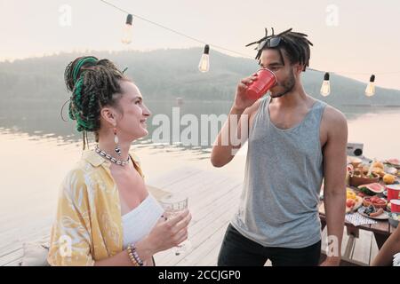 Junge Frau lächelt zu jungen Mann, während sie auf stehen Ein Pier im Freien und Cocktails trinken Stockfoto