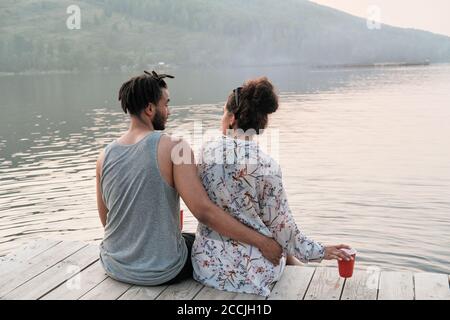 Rückansicht des jungen Paares umarmen und genießen die schöne Blick auf den See, während man auf einem Pier sitzt Stockfoto