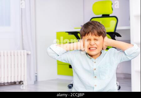 Autistischer Junge im Schulalter mit stark negativem Gesichtsausdruck, der sich schließt Ohren und grinst Zähne sitzen in seinem Zimmer Stockfoto