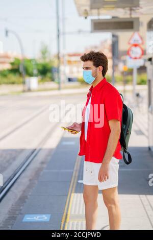 Junger Mann trägt eine chirurgische Maske während der Wartezeit auf ein Zug an einem Außenbahnhof Stockfoto