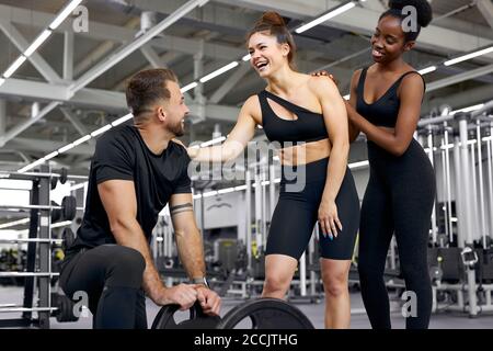 Entspannen Sie sich Zeit im Fitnessstudio, zwei verschiedene Frauen haben mit Sportler reden, sie diskutieren etwas, lächeln, Ruhe haben, eine Pause während des Trainings, Gewichtheben Stockfoto