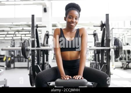 Portrait of adorable african Lady sitzend im Fitnessstudio, junge Dame in Sportbekleidung nach Crossfit-Übungen, posieren, Lächeln. Gesundes Lifestyle-Konzept Stockfoto