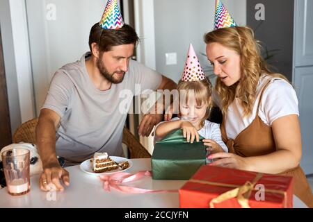 Kleine kaukasische Mädchen in Erwartung des Empfangs, Auspacken Geschenke von Eltern. Liebenswert glückliches Mädchen feiern Geburtstag zu Hause mit der Familie Stockfoto