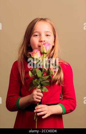 Junge hübsche 8-jährige Mädchen trägt ein rotes Kleid Riechende Blumen isoliert auf orangefarbenem Hintergrund Stockfoto