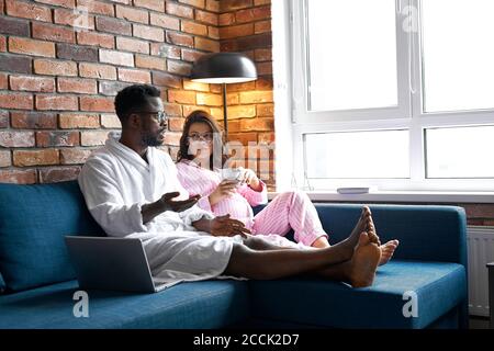 Glückliche multi-ethnische Familie haben Ruhe, zu Hause reden, Freizeit. Ehemann und schwangere Frau in modernen Zimmer-Interieur. Das Konzept der Schwangerschaft, Ruhe und ex Stockfoto