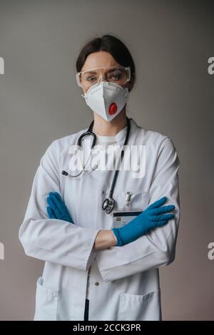 Ärztin trägt Maske und Schutzbrille mit gekreuzten Armen Im Krankenhaus gegen die Wand stehen Stockfoto