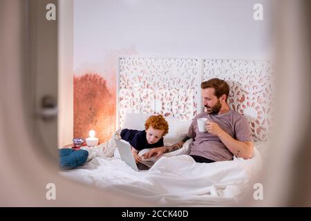 Junge, der sich den Laptop ansieht, der vom Vater auf dem Bett benutzt wird Stockfoto