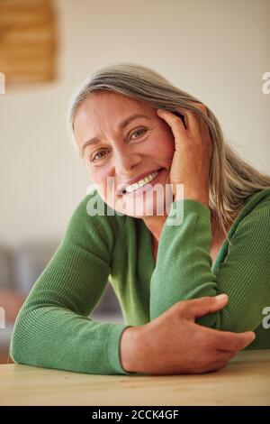 Glückliche Frau mit der Hand auf der Wange, die am Tisch sitzt Zu Hause Stockfoto