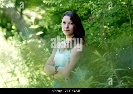 Junge Frau mit gekreuzten Armen, die inmitten von Pflanzen im Wald steht Stockfoto