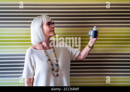 Stilvolle ältere Frau trägt eine Sonnenbrille und nimmt Selfie mit der Kamera entgegen Wand zu Hause Stockfoto