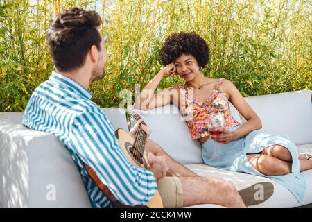 Frau hält Rotweinglas, während sie den Mann beim Spielen ansieht Gitarre auf dem Sofa im Penthouse Patio Stockfoto