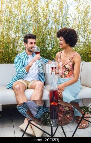 Glückliches multiethnisches Paar mit Rotwein, während es auf dem Sofa sitzt Im Penthouse Patio Stockfoto