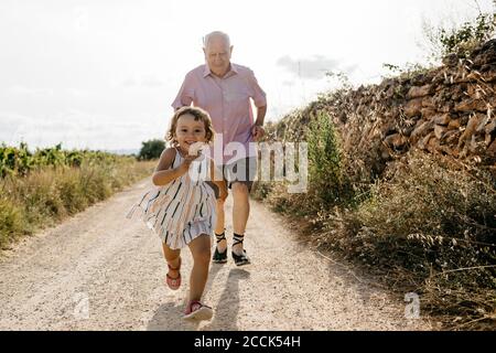 Großvater läuft hinter verspielte Enkelin auf Feldweg gegen den Himmel Stockfoto