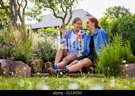 Lächelnde Freunde essen Wassermelone, während sie inmitten von Pflanzen im Garten sitzen Stockfoto