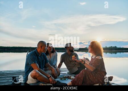 Freunde, die bei Sonnenuntergang am Steg an einem See picknicken Stockfoto