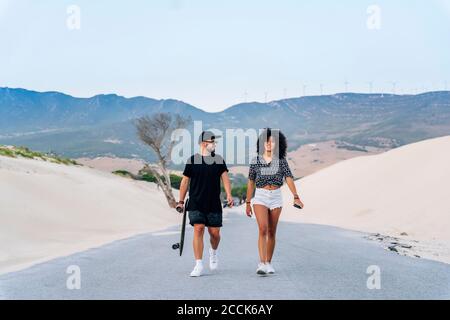 Lächelndes multiethnisches Paar, das auf der Straße zwischen Sanddünen gegen läuft Der Himmel ist klar Stockfoto