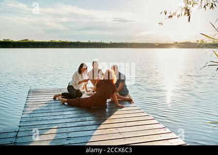 Freunde, die bei Sonnenuntergang am Steg an einem See picknicken Stockfoto