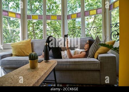 Frau liest Buch, während sie sich auf dem Sofa im Wohnzimmer entspannt Stockfoto