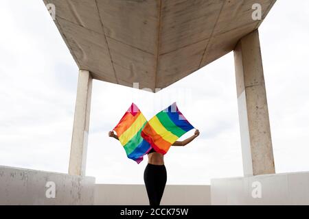 Mittlere Erwachsene Frau winkt Regenbogenfahnen, während sie auf gebaut steht Struktur gegen Himmel Stockfoto