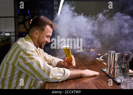 Fröhlicher junger Mann genießt Bier in der Bar allein, sitzt lächelnd, Spaß haben, nach einem harten Tag Stockfoto